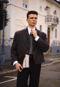 Siva krojena jakna preko sive veste, bijela košulja i tamne hlače. Drži smeškanu torbu i novine s tekstom u crnom na bijeloj pozadini.