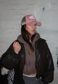 Hnedá kožená bomber bunda, šedá skrátená mikina s kapucňou, károvaná sukňa a ružová čiapka s priehľadným zadným dielom, pózujúca pred textúrovanou stenou.