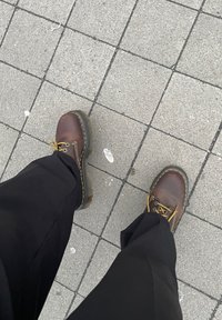 Rudos odiniai aulinukai iki kulkšnies su geltonomis siūlėmis ir apvaliais raišteliais, derinami su laisvais juodais kelnėmis, ant pilko tekstūruoto šaligatvio.