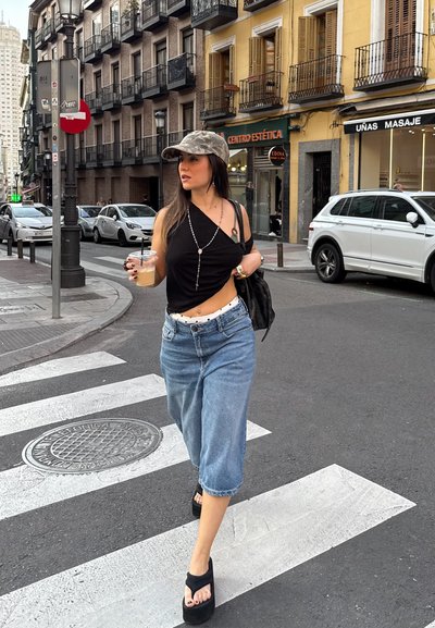 Top negro corto, jeans anchos de color azul claro, sandalias negras de plataforma, gorra de camuflaje gris; sosteniendo una bebida mientras camina por un paso de peatones.