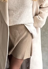 Jupe en laine beige avec un ourlet évasé, superposée sous un haut en tricot clair et un manteau beige doux. Texture subtile et tons neutres.