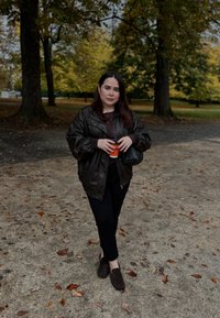 Schwarze Lederjacke, dunkles Oberteil, schwarze Hose, braune Schuhe. Hält eine Tasse und steht auf einem Weg mit gefallenem Laub und Bäumen im Hintergrund.