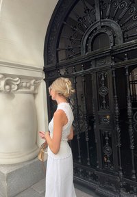Bílé, strukturované, zkrácené krajkové tričko s prolamovanými okraji; kombinované s volnou bílou sukní. Pohled zezadu blízko zdobených černých železných bran.