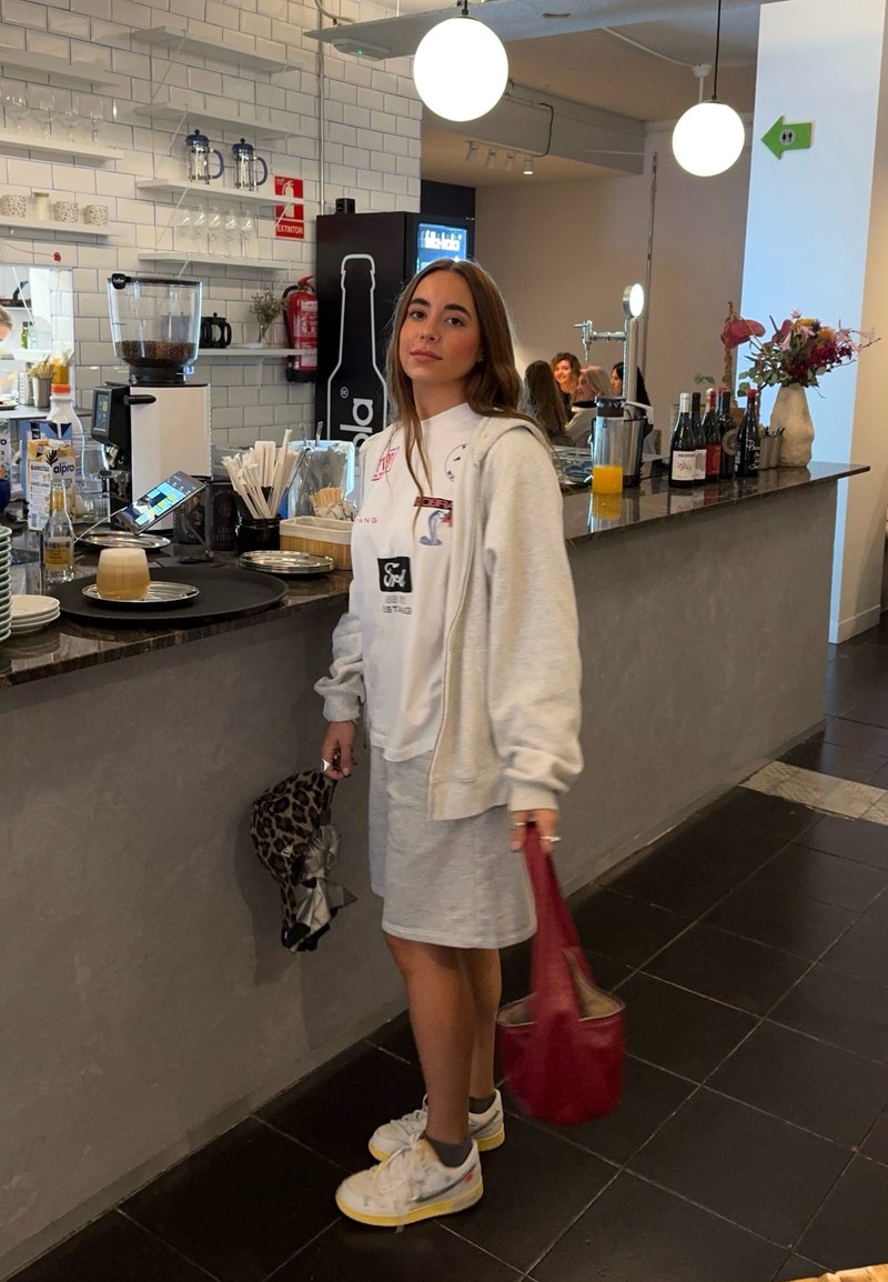 Junge Frau in lässiger Kleidung, die eine rote Tasche und einen Schal mit Leopardenmuster hält, steht an der CafTheke mit Getränken und Utensilien.