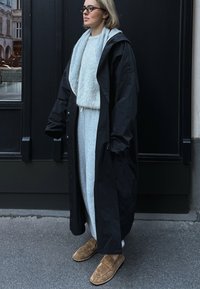 Fekete hosszú kabát kapucnival, szürke pulóver és azonos színű nadrág felett. Barna velúr slip-on cipők egészítik ki az öltözetet. Szemüveg viselve.