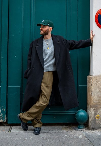 Abrigo largo negro, suéter gris, pantalones oliva y zapatos negros. Gorra de béisbol verde y collar con colgante. Frente a una puerta color verde azulado.