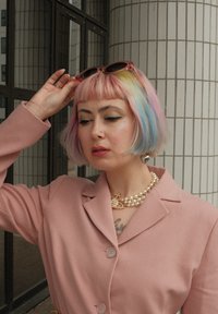 Lyserød skreddersydd blazer med fire knapper, lagdelt halskjede av falske perler, pastell regnbuehår, og runde solbriller hvilende på pannen.