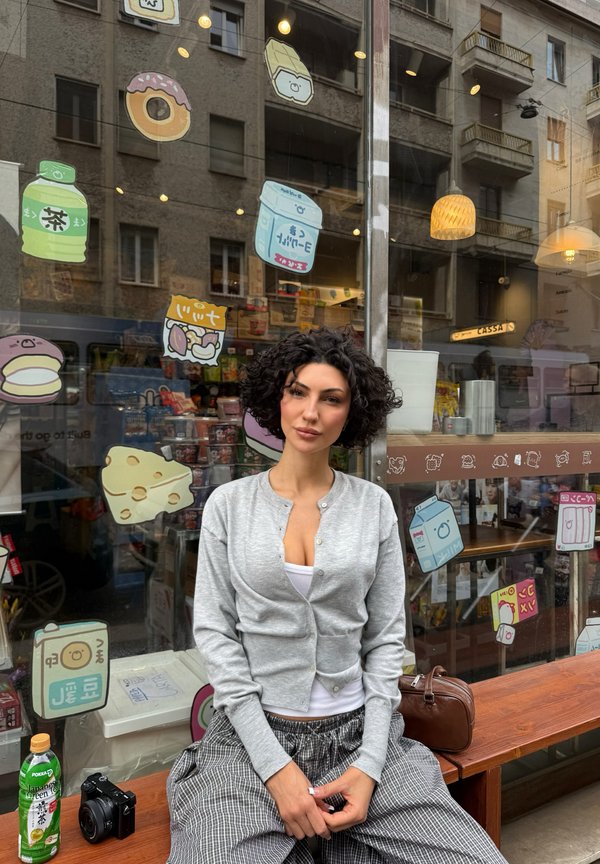 Femme aux cheveux bouclés assise devant un magasin avec des illustrations de nourriture sur la vitrine, portant un cardigan gris et un pantalon à carreaux.