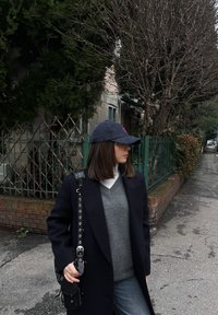 Γυναίκα ντυμένη με σκούρο παλτό, γκρι πουλόβερ και σκουφάκι να περπατά σε πεζοδρόμιο δίπλα σε πράσινο φράχτη και γυμνά δέντρα σε μια συννεφιασμένη μέρα.