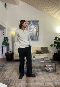 Jasnoszara koszula henley z długim rękawem i trzema guzikami, zestawiona z czarnymi, luźnymi spodniami i czarnym obuwiem. Nowoczesne, wewnętrzne wnętrze.