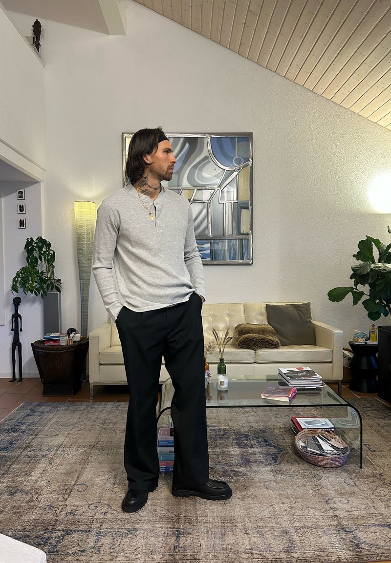 Chemise henley à manches longues gris clair avec patte de boutonnage à trois boutons, associée à un pantalon ample noir et des chaussures noires. Cadre intérieur moderne.