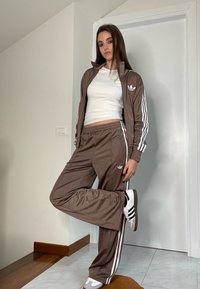 Rudas treniruoklių komplektas su baltais dryžiais, derančios kelnės ir striukė, taip pat baltos sportinės bateliai su juodais akcentais. Lygus, blizgus paviršius.