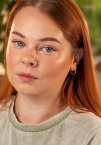 Portret osoby z długimi, rudymi włosami i niebieskimi oczami. Ma na sobie jasnoniebieską koszulę z krótkim rękawem. Charakterystyczne cechy to kolczyk w nosie i okrągłe kolczyki w uszach.