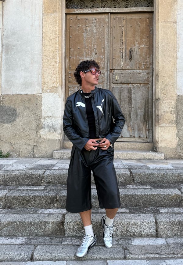 Giubbotto bomber in pelle nera con logo bianco, abbinato a pantaloni corti in pelle nera. Scarpe sportive argentate e accessori con catena argentata. Sfondo con gradini in pietra.