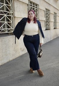 Farmerdzseki és magasderekú sötét farmer, krémszínű pulóverrel és barna bebújós cipővel párosítva; egy kis barna táskát tart a kezében.