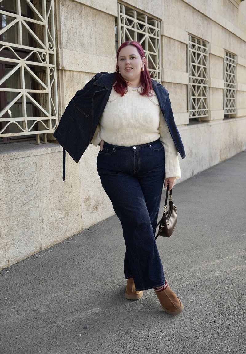 Denim jacket and high-waisted dark jeans paired with a cream sweater and tan slip-on shoes; holds a small brown bag.