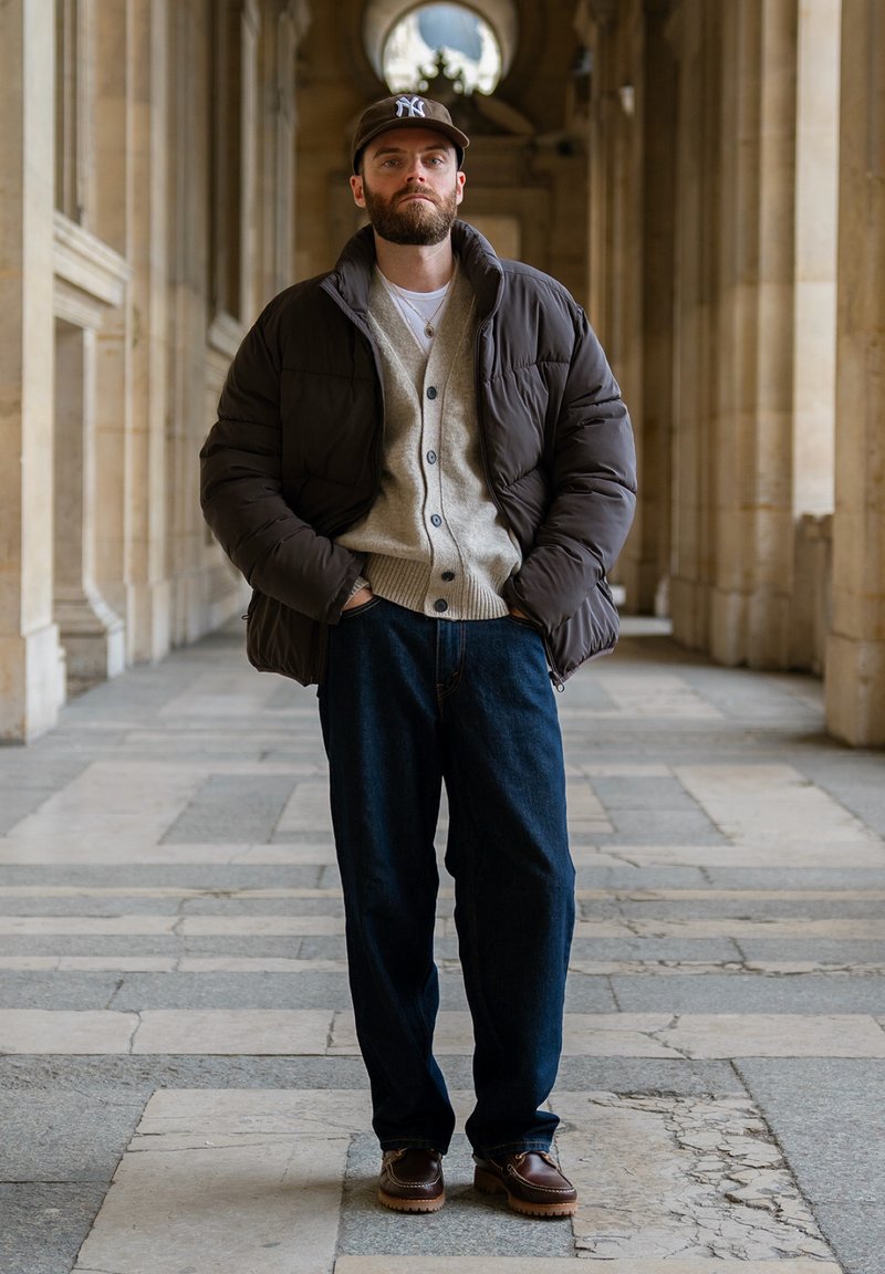 Homme barbu portant une doudoune marron, un cardigan beige, un jean bleu et une casquette de baseball, debout dans un couloir voûté en pierre, les mains dans les poches.