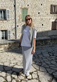 Grijze lange rok gecombineerd met een wit t-shirt en een grijze cardigan. Draag het met witte sneakers. Gelaagd stenen achtergrond met groene luiken.