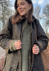 Hnědá voskovaná bavlněná bunda na zip, pod ní zelený pletený svetr a na rukávech kostkované detaily. Obsahuje boční tašku a neutrální pozadí.