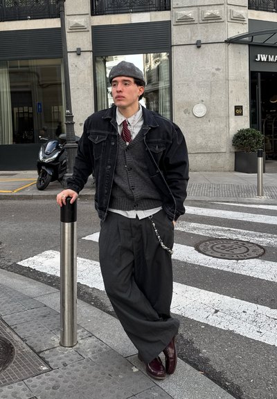 Chaqueta de mezclilla negra, chaleco de punto gris, camisa blanca a rayas, corbata burdeos, pantalones anchos grises, zapatos burdeos pulidos, detalle de cadena en la cintura.