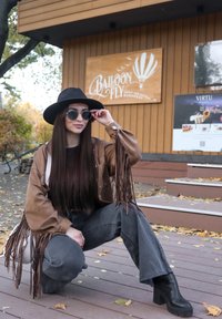 Bruine jas met franjes, zwarte hoed met brede rand, ronde zwarte zonnebril, grijze wijde jeans en zwarte laarzen met hak. Houten achtergrond met borden.