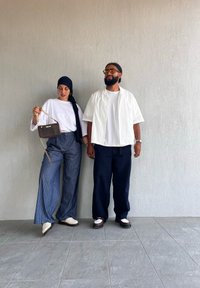 Couple se tenant la main ; femme portant une chemise blanche oversize, un pantalon large rayé et tenant un petit sac à main ; homme en chemise blanche et pantalon marine.