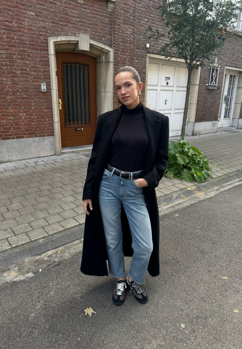 Jeune femme portant un long manteau noir, un pull à col roulé noir, un jean bleu clair et des baskets se tient dans une rue urbaine près de bâtiments en briques et d'un arbre.