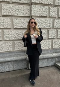 Crni oversized blazer, bijeli rebrasti krojeni top, crna duga suknja i crne tenisice s bijelim prugama. Drži prozirnu čašu s slamkom.