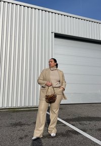 Μπεζ συνθετικό δερμάτινο κοστούμι με φαρδιές παντελόνες και ταιριαστό blazer. Ανοιχτό μπεζ ζιβάγκο από κάτω. Καφέ τσάντα με σχέδιο και αλυσίδα.