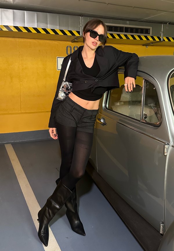 Chaqueta negra corta sobre una blusa negra, pantalones cortos de rayas, medias negras transparentes y botas negras hasta la rodilla. Un bolso transparente y gafas de sol añaden detalle.