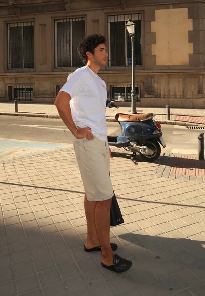 Conjunto casual que incluye una camisa blanca de manga corta, pantalones cortos beige, zapatos negros sin cordones y un bolso negro. De pie en una calle pavimentada al lado de un patinete.