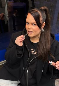 En person med langt hår iført en sort oversized jakke påfører læbepomade med en sølvfarvet og gennemsigtig tube, mens de sidder foran en blå baggrund.