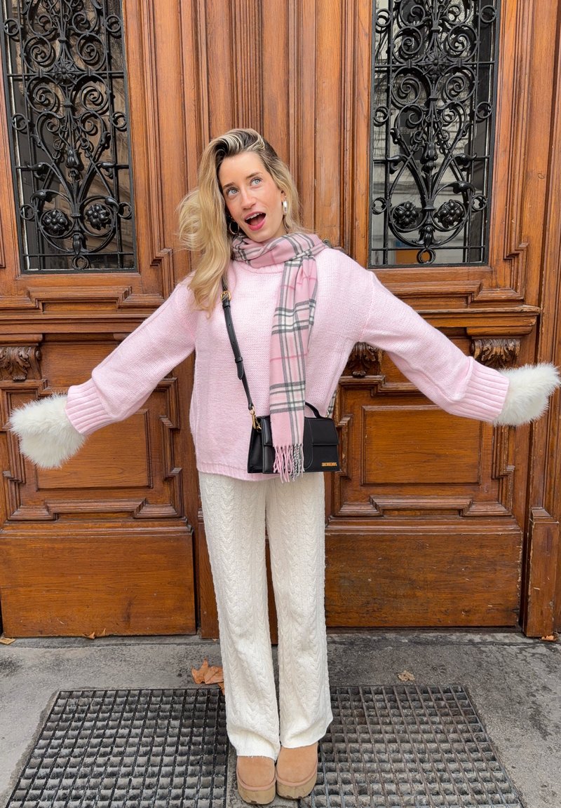Pull en maille rose avec des poignets en fourrure blanche, pantalon blanc texturé et écharpe à carreaux rose. Sac bandoulière noir et bottes beige complètent la tenue.