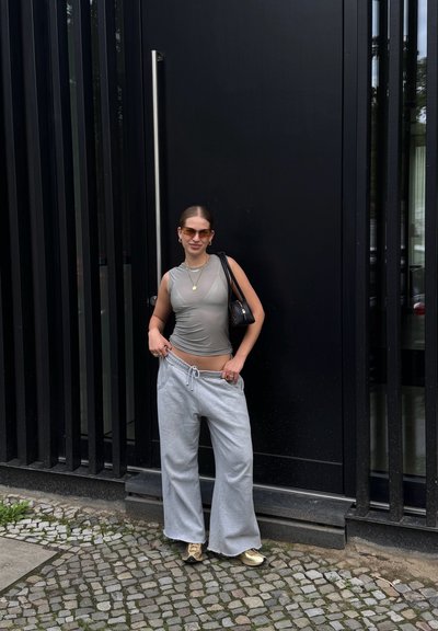 Pantalones de chándal amplios grises, camiseta ajustada gris, gafas de sol negras, collares en capas y un bolso negro, posando frente a una pared vertical de tablones negros.