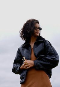 Zwarte leren jas met rits en bontkraag; bruine jurk eronder; houdt een metalen clutch vast; draagt kleine zwarte zonnebrillen.