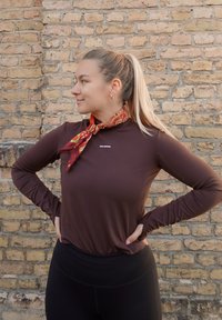 Barna, hosszú ujjú ing magas gallérral, amelyen egy diszkrét logó található. Egy mintás piros és narancssárga sál díszíti a nyakat.