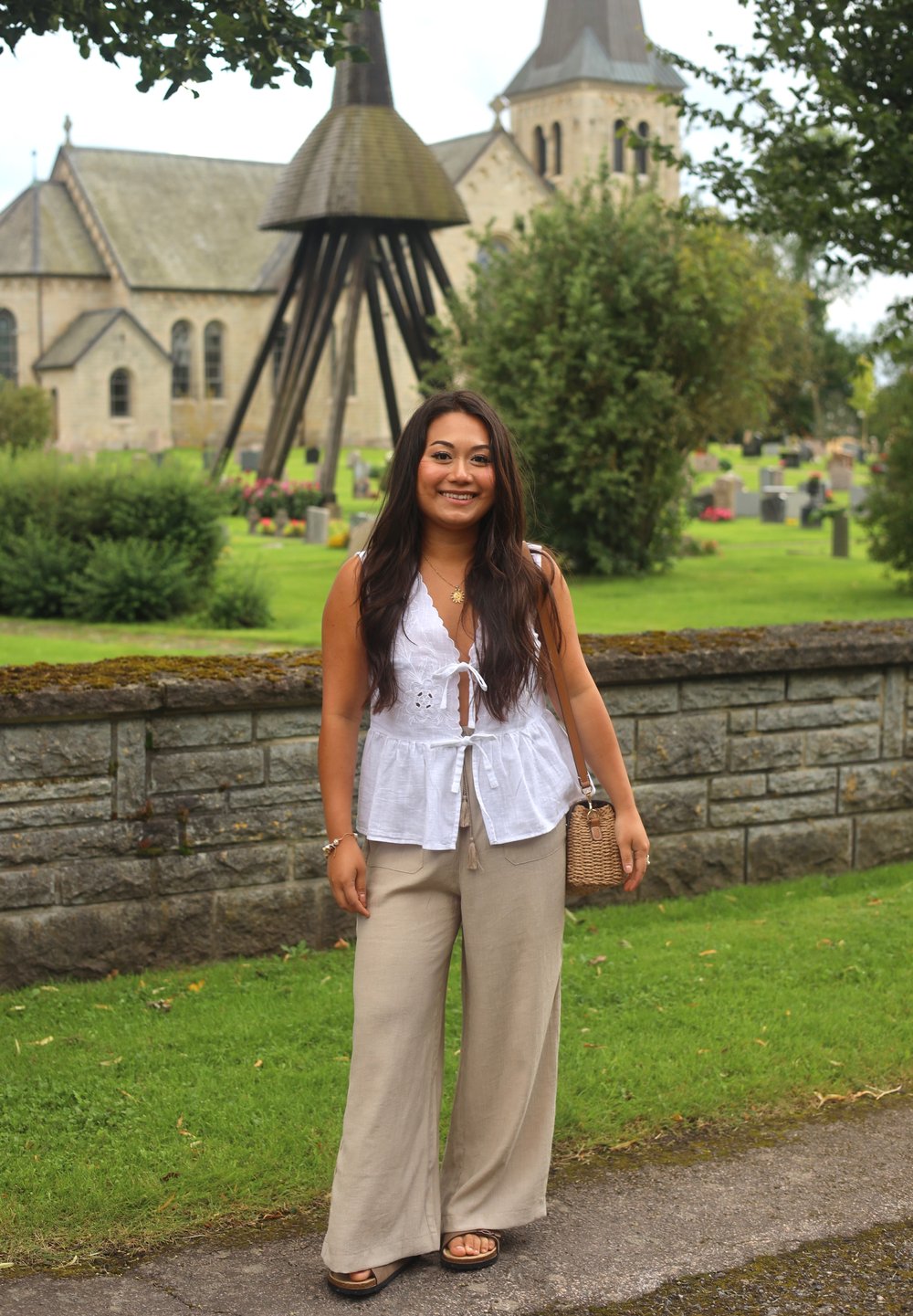 Valge lina topp nööpdetailiga, beežid laiad püksid ja mustad sandaalid. Hoides kootud kotti, seisab rohelises pargis kiviseina lähedal.