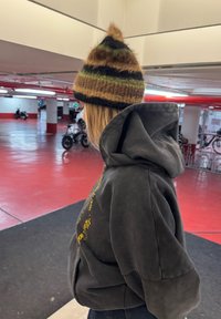 Berretto lavorato a maglia in marrone, verde e nero con cima appuntita. Felpa con cappuccio grigio scuro con tasca frontale e vestibilità comoda. Sfondo di un parcheggio coperto.