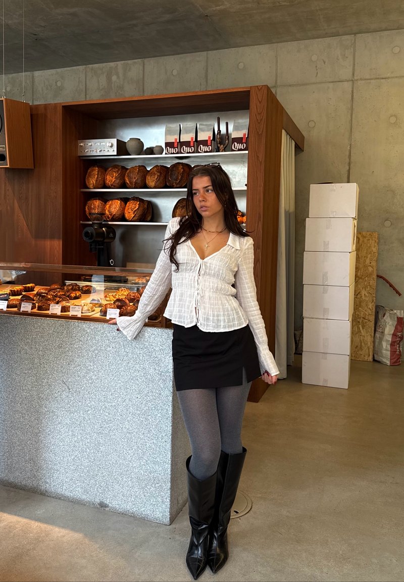 Camisa branca de botão com tecido texturizado, mini saia preta, collants cinzentos e botas altas pretas com bico fino. Vitrine de padaria ao fundo.