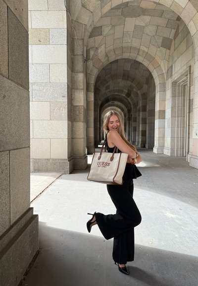 Bolsa tote de lona beige con detalles de cuero marrón, con la inscripción "GUESS EST 1981" en color burdeos. Modelo viste ropa negra y zapatos de tacón.