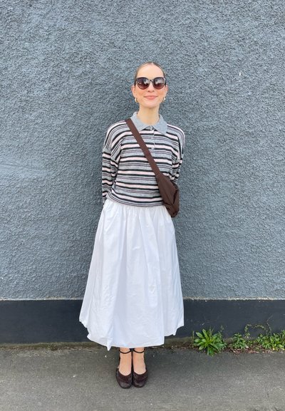 Mujer con polo de manga larga a rayas, falda blanca, gafas de sol oscuras y bolso bandolera marrón, de pie contra una pared gris texturizada.