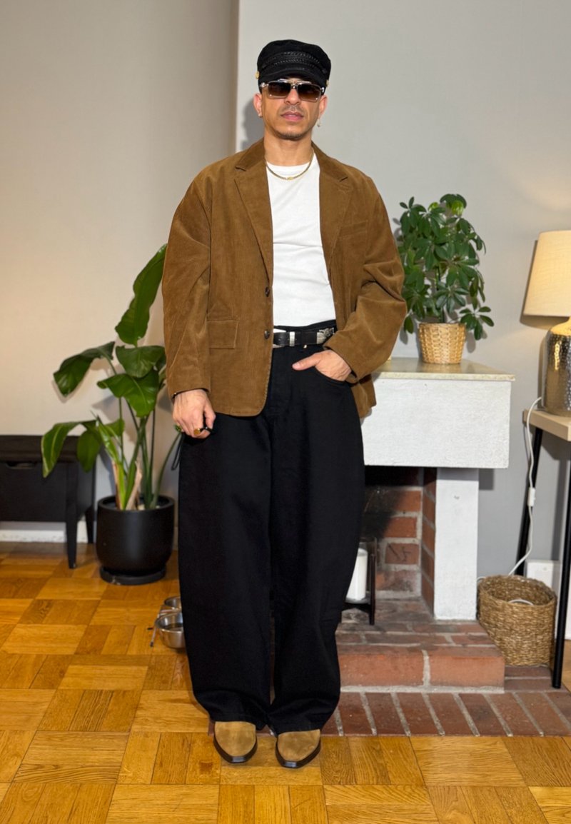 Mand iført en brun blazer, hvid skjorte, sorte vide bukser, brune sko, sort kasket og solbriller, står indendørs nær planter og en pejs.