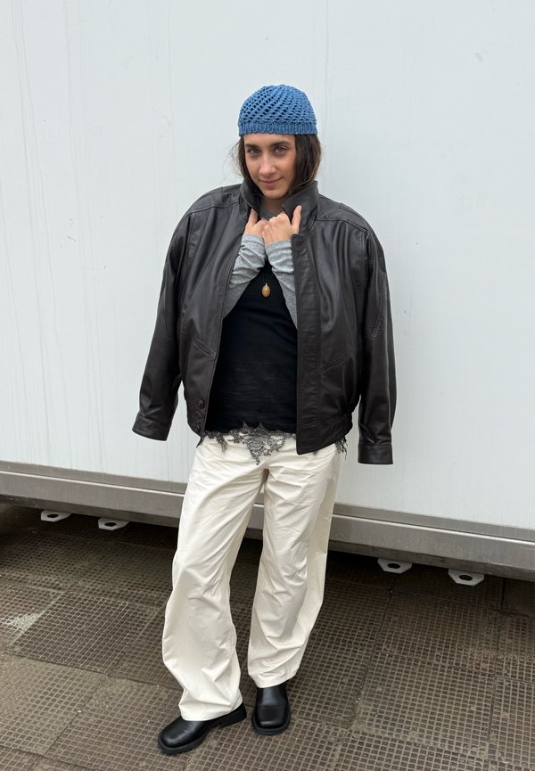 Osoba nosząca niebieską dzianą czapkę, czarną skórzaną kurtkę na czarnych i szarych warstwowych bluzkach, białe spodnie i czarne buty, stojąca na kafelkowej powierzchni.