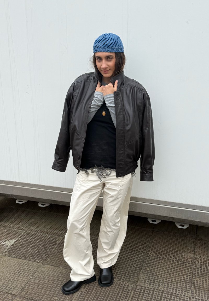 Persona che indossa un berretto blu di maglia, giacca di pelle nera sopra top neri e grigi a strati, pantaloni bianchi e stivali neri, in piedi su una superficie piastrellata.