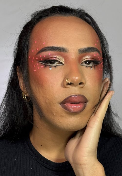 Primo piano di un volto con trucco che presenta puntini rosa e accenti scintillanti. La pelle appare liscia; i capelli sono lunghi e scuri. È visibile una maglietta nera.