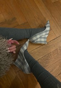 Pieds en collants gris côtelés portant des escarpins à bouts pointus gris et blancs à carreaux sur un sol en bois, avec une main et une manche duveteuse partiellement visibles.