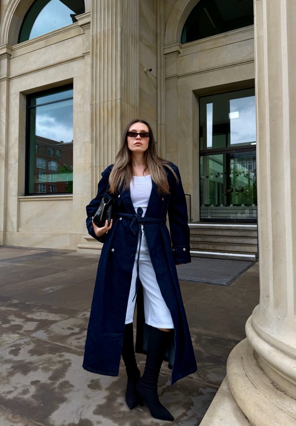 Femme portant un long manteau sombre, pantalon blanc, bottes noires, lunettes de soleil, tenant un sac noir, debout devant un bâtiment en pierre avec des colonnes.