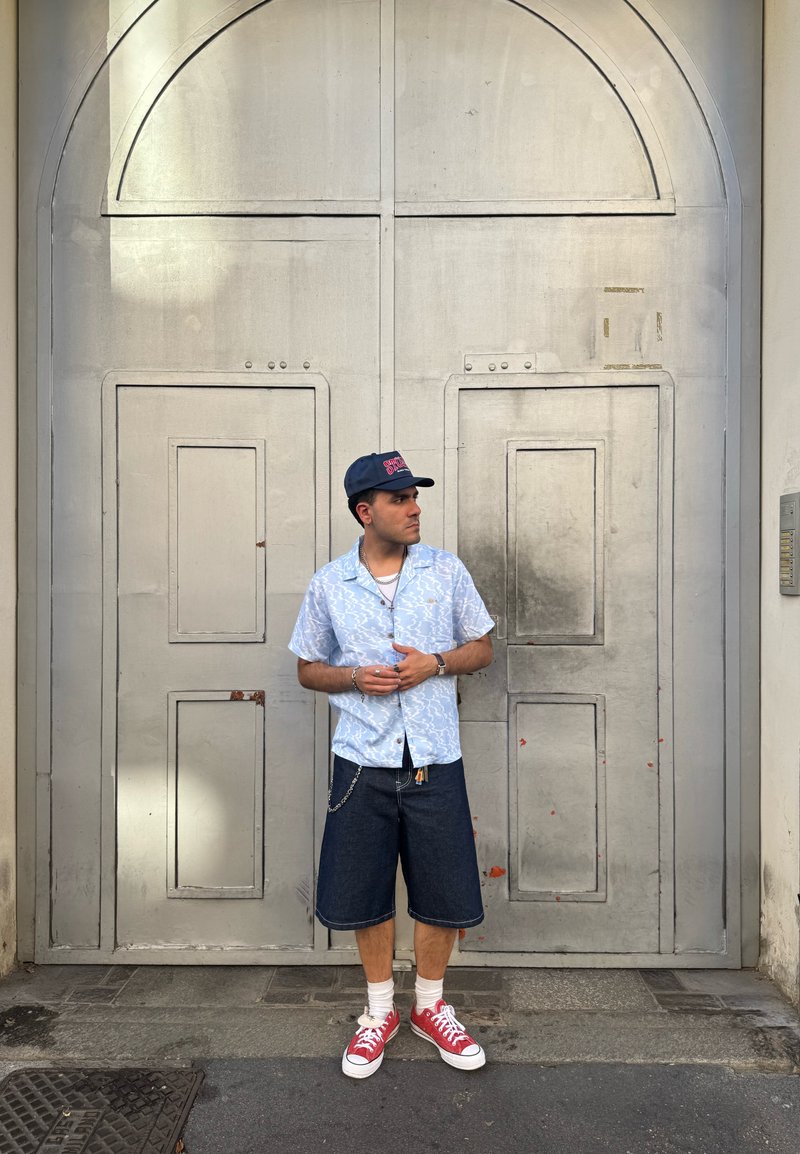 Chemise à manches courtes, de couleur bleu clair avec un motif, short en denim, baskets rouges, détail en chaîne argentée, posé devant une porte grise usée.