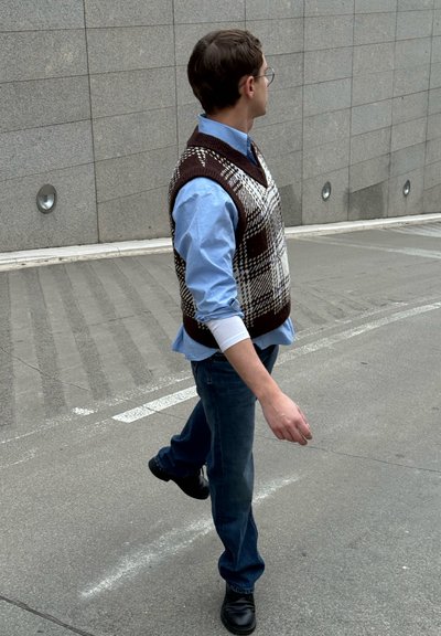 Chaleco de punto a cuadros marrón y blanco sobre una camisa azul claro, combinado con pantalones vaqueros azules y zapatos negros, llevado por una persona caminando.
