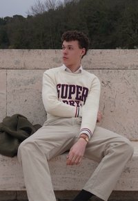 Pull en tricot crème avec le logo "SUPER", pantalon beige et une montre-bracelet brune, assis sur une surface en pierre avec une veste verte à côté.